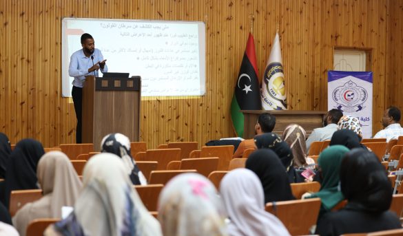 Lecturer at a podium presenting to an audience in a wooden-floored lecture hall with flags and a projection screen displaying Arabic text.