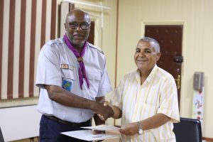 Two men shake hands while exchanging papers indoors; one wears a scout uniform with a purple neckerchief, the other a striped shirt and watch.