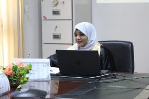 Woman in a light gray hijab sits at a desk with an HP laptop, flowers on the left, and an office setting in the background.