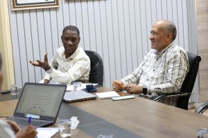Two men seated at a conference table in discussion, one gesturing with his hand while the other looks on, with laptops and notebooks in front of them.