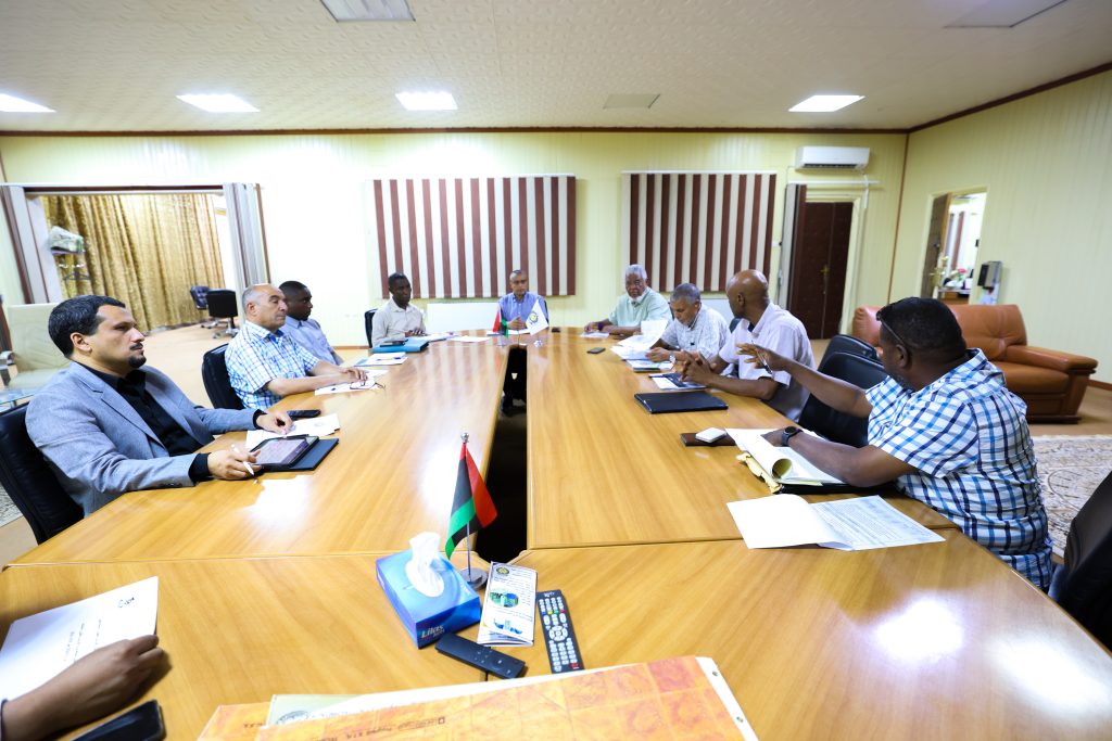 Business meeting in a bright conference room with several people seated around a long wooden table and flags in the center.