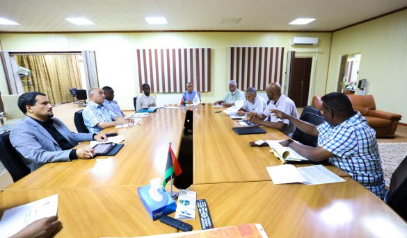 EB204D0B-B2F5-4CC1-B791-A4ED124A4BB0 - جامعة سبها | Sebha University Business meeting in a bright conference room with several people seated around a long wooden table and flags in the center.