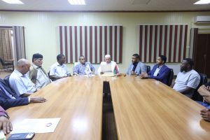 EC18ECEB-D3E7-45A6-A910-4FE2711084A7 - جامعة سبها | Sebha University Group of men in business attire seated around a long wooden conference table in a meeting room with striped blinds behind them.