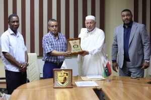 EDF31BC3-DF4E-43FA-8A1A-87754EDEE1E9 - جامعة سبها | Sebha University Four men in a formal meeting: one presents a plaque to a senior man in white robes as others look on, in a conference room with striped wall panels and a table with documents and a small flag.