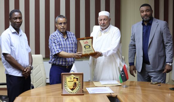 EDF31BC3-DF4E-43FA-8A1A-87754EDEE1E9 - جامعة سبها | Sebha University Four men in a formal meeting: one presents a plaque to a senior man in white robes as others look on, in a conference room with striped wall panels and a table with documents and a small flag.
