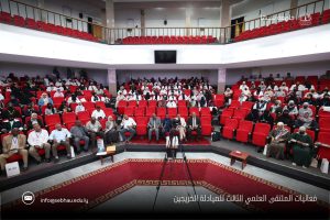 Auditorium full of attendees in red seats, with a camera tripod centered at the front; Arabic watermark text at bottom right.