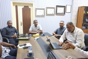 F07A653E-9047-493E-B1DE-20A8FE62D955 - جامعة سبها | Sebha University Four men sit around a conference table in a room with framed certificates on the wall, laptops and cups in front of them, engaged in a meeting.