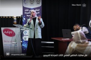 Woman wearing hijab speaks into a microphone at a clear acrylic podium on a stage during a conference, with a blue banner and a man at a laptop nearby.