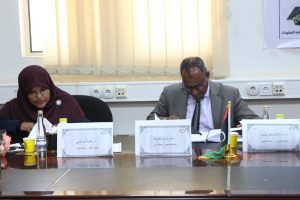 Two people seated at a conference table signing papers, with name placards in Arabic in front of them and bottled drinks nearby.