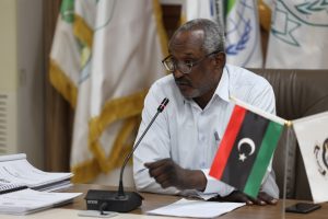 Man with glasses speaking into a microphone at a formal meeting, seated at a desk with papers and flags in the background, Libyan flag visible on the table