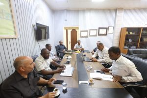 A business meeting in a conference room with eight men seated around a long table, laptops, papers and coffee cups nearby.