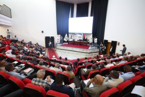 F9D061F7-F65F-4737-BE83-6578F5B8A8C6 - جامعة سبها | Sebha University Audience seated in red theater chairs watching a conference on a stage with banners and a large screen at the front.