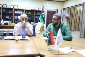 Two men sit at a conference table in a sunny office, attentively discussing with notebooks and flags in view behind them.