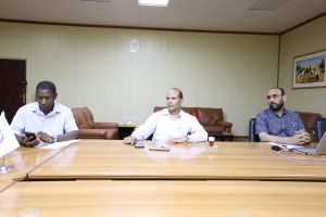 Three men sit at a long wooden conference table in a beige meeting room; left man uses a smartphone, center man looks ahead, right man in a checked shirt sits with hands folded; camel painting on wall.