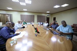 People seated around a large oval wooden conference table, discussing documents with laptops and cups in a meeting room.