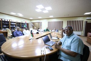 Group of men seated around a curved conference table with laptops, Libyan flags, and awards on shelves behind them.