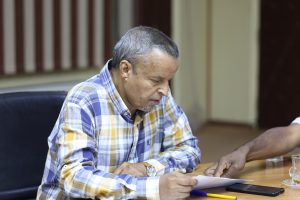 Older man in a blue-yellow plaid shirt sits at a table, looking at a document he’s reviewing at a meeting.