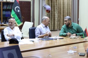 Three men sit around a wooden conference table in an office, engaged in conversation, with a Libyan flag in the background.