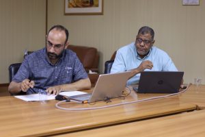 Two men sit at a conference table with laptops and papers, discussing work in a office setting.”,