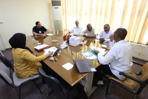 A diverse group of six people seated around a large conference table in a bright room, discussing documents.