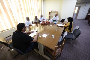 A group of six people sit around a conference table reviewing papers, with a woman in a mustard outfit and black hijab on the right end of the table, and a laptop nearby.