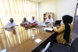 Five professionals sit around a glass conference table reviewing documents, with a floral centerpiece and a laptop nearby. A woman in a hijab sits on the right.