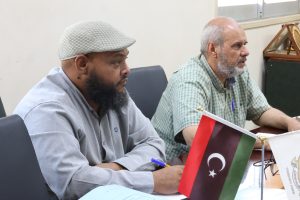 WhatsApp Image 2026-06-02 at 12.45.50 PM (2) - جامعة سبها | Sebha University Two men sit at a conference table, one wearing a light knit cap and gray shirt, the other in a green checkered shirt, with Libyan flags on the table in front of them.