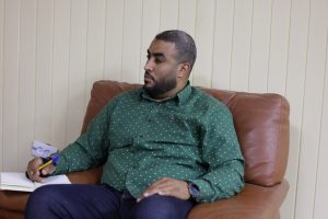 Man with a beard sits on a brown leather couch, wearing a green patterned shirt, writing with a yellow pencil on papers in front of him.
