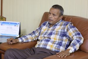 Older man with gray hair sits on a brown leather sofa in a plaid shirt, gazing to the side in a casual office setting, with a framed certificate on a desk nearby.