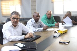 Four men sit at a conference table in a meeting room, focused on documents and laptops with papers and notebooks scattered on the table.