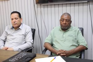 Two men seated at a conference table in a room with vertical metal panel walls; left man in gray shirt, right man in green polo.