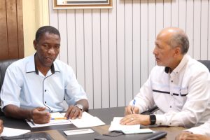 Two men sit at a table signing papers in an office setting, pens in hand and documents spread out before them.