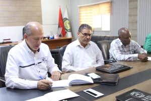 Three men sit at a conference table reviewing documents, one writing in a notebook while the others read or think.