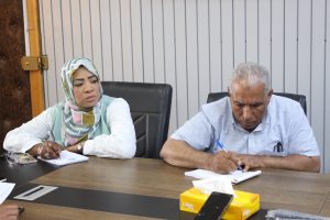 Two colleagues sit at a conference table, the woman in a patterned scarf appears to be listening while writing on paper, and the man beside her is focused on his own notes.