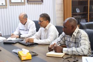 Three men in a meeting around a conference table, reviewing and signing documents.