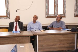 Three men in business attire seated at a long conference table, listening during a meeting, with notebooks and pens in front.