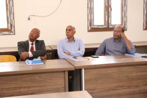 Three men sit at a long wooden conference table in a meeting room, listening attentively.