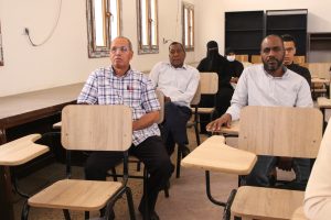 Group of adults seated in a classroom; man in a plaid shirt foreground with others behind him in rows of desks.