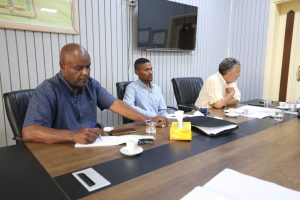 Three men sit at a conference table reviewing documents in a meeting room with a TV on the wall behind them.
