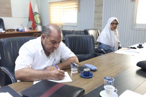 Man in a white shirt writes on papers at a large conference table; a woman in a hijab sits in the background.