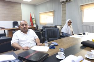 Man in a white shirt sits at a conference table in a meeting room, papers and cups scattered nearby, woman in hijab in the background.] ,