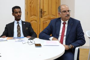 WhatsApp Image 2026-06-10 at 4.54.06 PM (1) - جامعة سبها | Sebha University Two men in suits sit at a white round table with documents; one looks to the side while the other stares forward, in a room with a wooden door behind them.