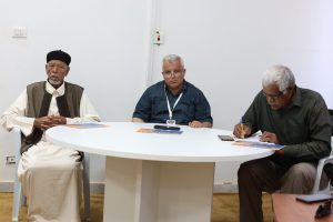 WhatsApp Image 2026-06-10 at 4.54.06 PM (3) - جامعة سبها | Sebha University Three men sit at a white round table in a plain room: the left man wears traditional clothing and a black hat, the center man has glasses and a lanyard, and the right man writes notes.