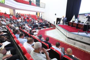 Large conference hall full of attendees in red seats, a speaker at a podium on stage with banners and cameras nearby.