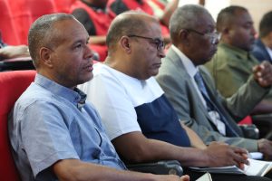 Four men seated in red auditorium chairs, attentively listening to a presentation at a conference setting.
