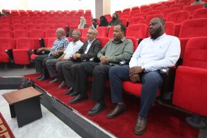 WhatsApp Image 2026-06-14 at 12.43.19 PM - جامعة سبها | Sebha University Six men sit in the front row of a red-upholstered auditorium, facing a stage area to the left.