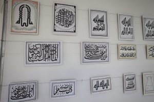 WhatsApp Image 2026-06-14 at 12.43.20 PM (2) - جامعة سبها | Sebha University Series of framed Arabic calligraphy artworks in black ink on white panels hung on a pale wall, forming a gallery display.