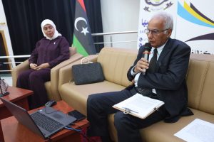 WhatsApp Image 2026-06-14 at 12.43.20 PM - جامعة سبها | Sebha University A man in a suit speaks into a handheld microphone while seated on a beige couch, with a woman in a hijab beside him and a laptop on a wooden table nearby.