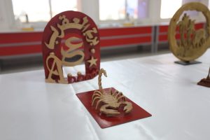 WhatsApp Image 2026-06-14 at 12.43.21 PM (1) - جامعة سبها | Sebha University Red and gold decorative arch sculpture on a white table at an exhibit, with a blurred workshop background.