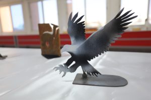 WhatsApp Image 2026-06-14 at 12.43.21 PM (3) - جامعة سبها | Sebha University Close-up of a dark metal bird sculpture with jagged, outstretched wings on a square pedestal in a bright gallery space.